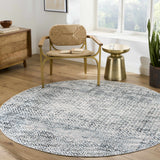 Living room scene with a round Askeaton Washable Area Rug in blue and gray, styled with a chair, side table, and wood dresser. Roomscene-Round