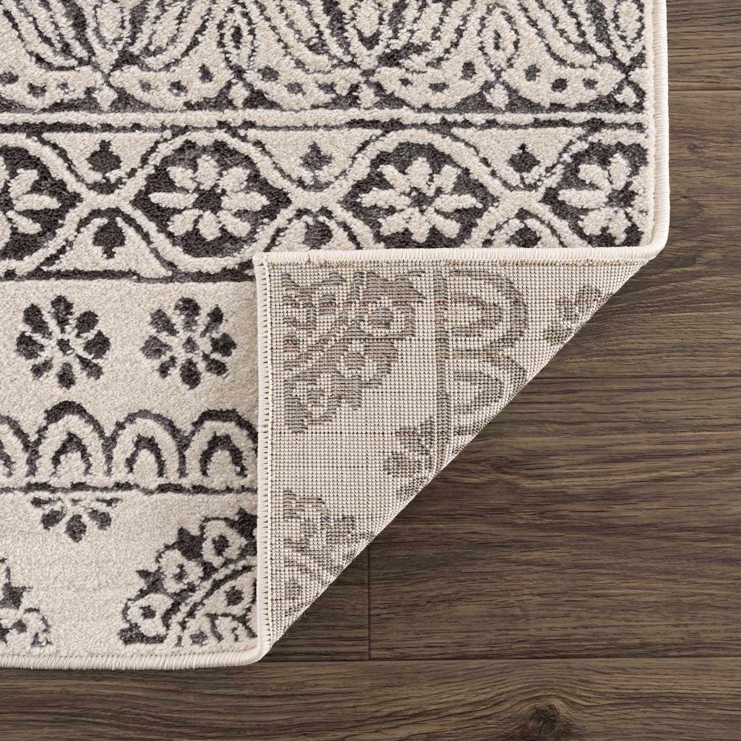 Close-up of the Warroad Area Rug showing a charcoal and light beige floral pattern and the rug's backing. Fold