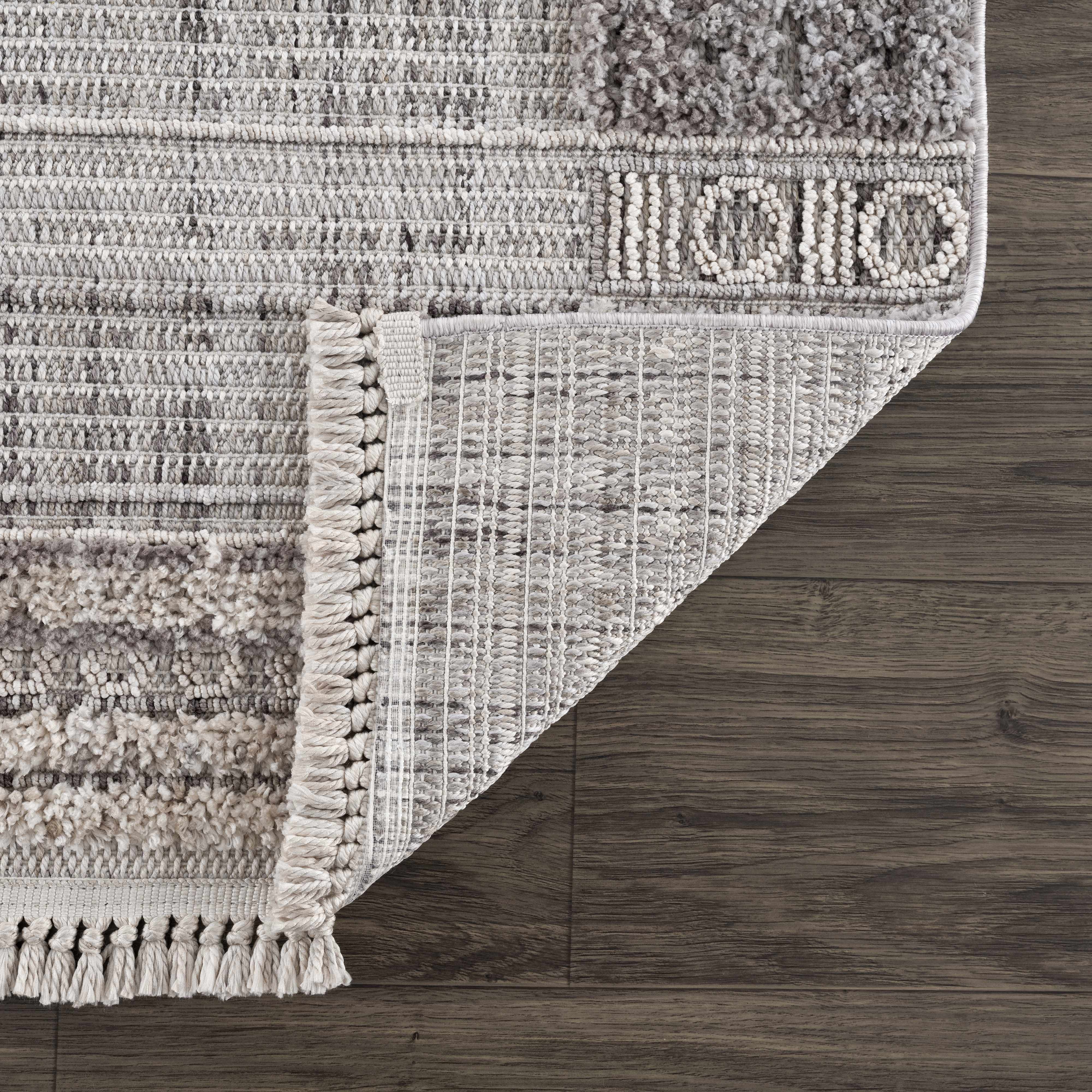 Close-up of Naivasha Elegant High/Low Area Rug showing charcoal, cream, and gray tones, plus fringe detail and rug backing. Fold
