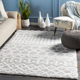 Living room scene with a light gray Vernonburg Shag Area Rug, beige sofa, patterned chair, and gray pouf on a dark wood floor. Roomscene-Rectangle